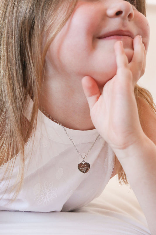 A close-up wedding lifestyle shot of a young girl resting her head on her hand, wearing a silver chain necklace with a heart-shaped pendant engraved with 'OUR LITTLE FLOWER GIRL'.
