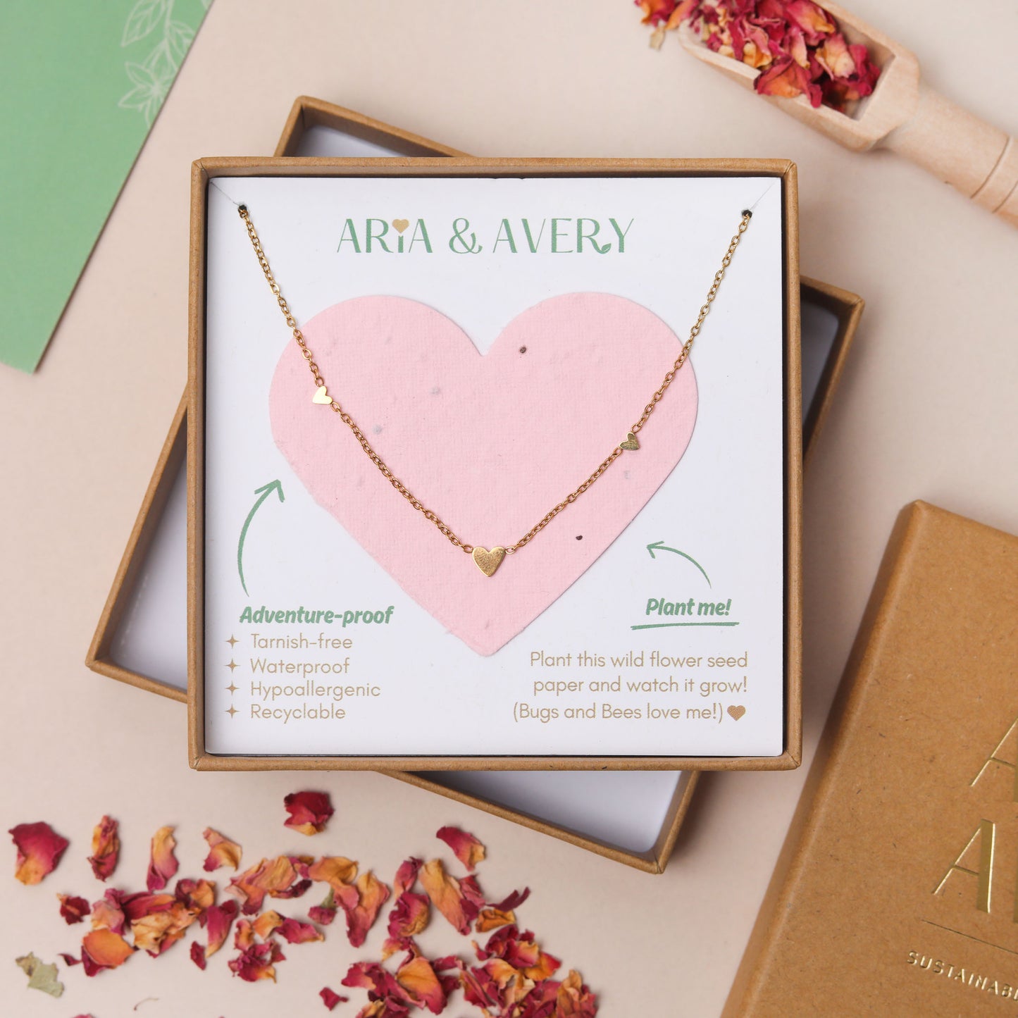 A gold heart necklace displayed on a pink plantable seed paper heart insert inside a gift box labeled ARIA & AVERY, styled on a beige surface with scattered dried rose petals and a green card.
