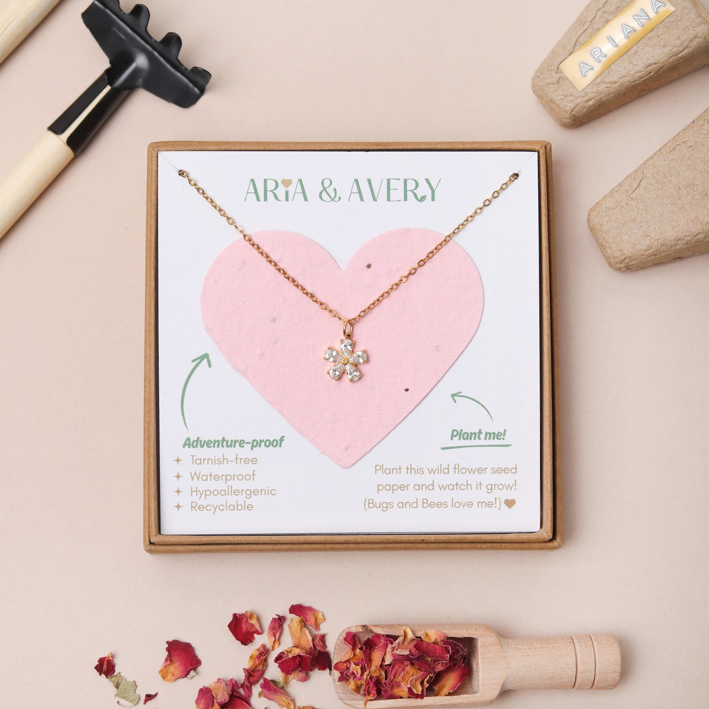 A gold-toned flower pendant necklace displayed on a pink heart-shaped seed paper backing card inside a square gift box. The card shows the brand name ARIA & AVERY and includes product feature text.