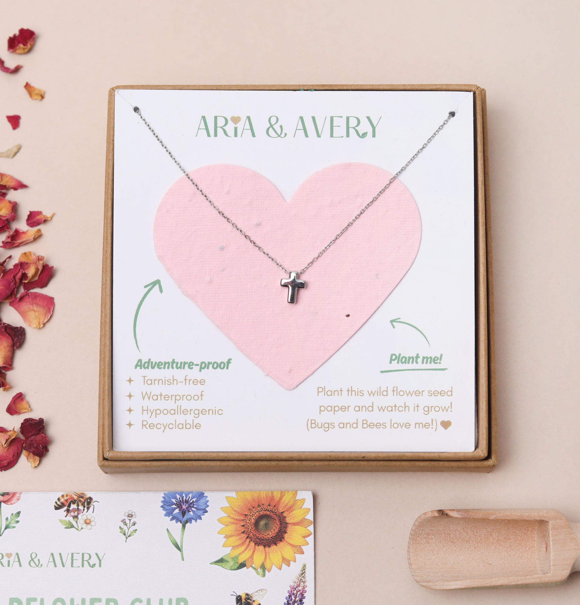 A children's silver cross pendant necklace displayed on a plantable seed paper heart-shaped card inside a kraft gift box, with the brand name ARIA & AVERY printed at the top. The card lists product features and includes a small card at the bottom with floral illustrations and dried flower petals on a beige background.