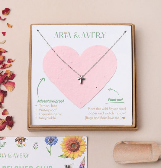 A children's silver cross pendant necklace displayed on a plantable seed paper heart-shaped card inside a kraft gift box, with the brand name ARIA & AVERY printed at the top. The card lists product features and includes a small card at the bottom with floral illustrations and dried flower petals on a beige background.