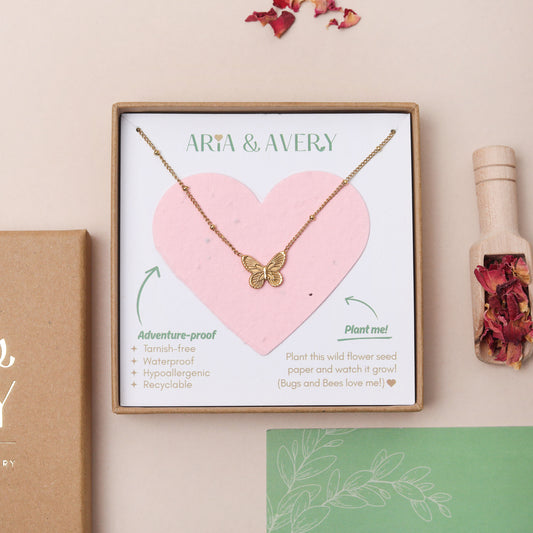 A gold butterfly necklace displayed on a pink heart-shaped seed paper card inside a square gift box labeled 'ARIA & AVERY'. The box is styled on a beige surface with a brown box edge, dried flowers, and a small bottle of dried petals nearby.