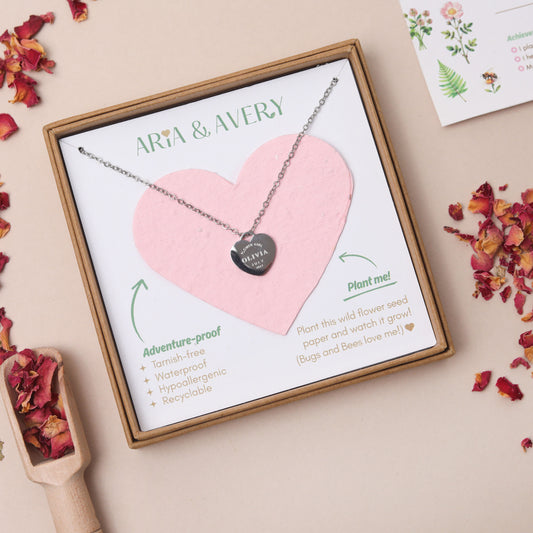 A personalised childrens heart-shaped silver pendant necklace with the words 'flower girl' engraved on a heart, with the flower girls name and wedding date engraved, presented in a gift box with a plantable seed paper pink heart insert and printed card. The brand name 'ARIA & AVERY' is visible at the top of the card.
